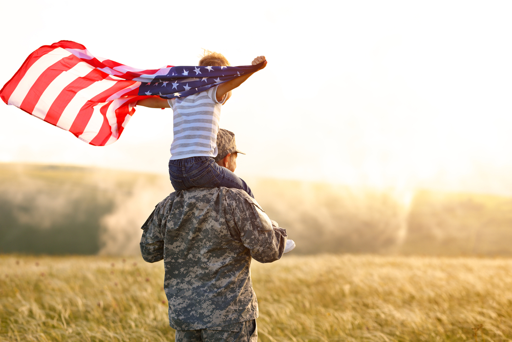 man in military uniform with kid on shoulders holding American flag